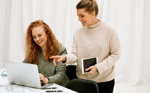Två glada kvinnliga entreprenadingenjörer står vid ett skrivbord. Den ena tjejen skriver på en dator och den andra håller i en ipad.
