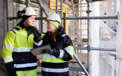 Bild på en man och en kvinna i varselkläder och bygghjälm på en byggarbetsplats som diskuterar med varandra
