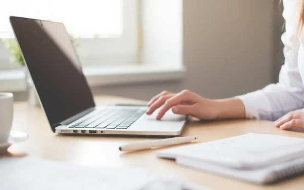 En kvinna sitter vid ett skrivbord och skriver på en laptop. Bredvid datorn finna en penna, anteckningsblock och en kopp med kaffe.