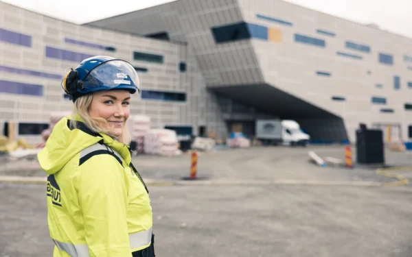 En kvinna står med reflexgula arbetskläder och skyddshjälm på sig, ute på en byggarbetsplats.