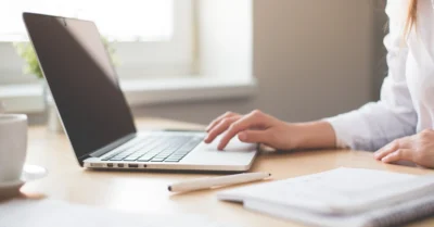 Detaljbild på en laptop på ett skrivbord, bilden är skuren så endast en hand syns arbeta på datorn, framför datorn ligger ett block och en penna.