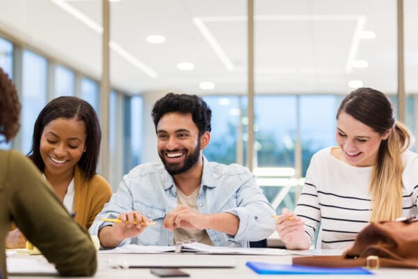 Tre glada studerande sitter vid ett bord i en skolmiljö.