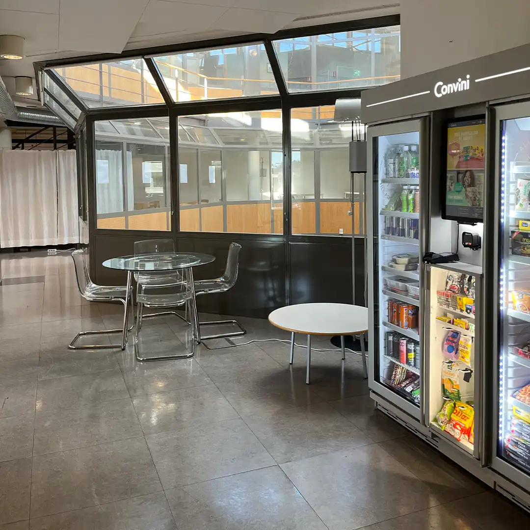 En hall med stora glaspartier och mycket ljusinsläpp. Här finns runda bord och en varuautomat.