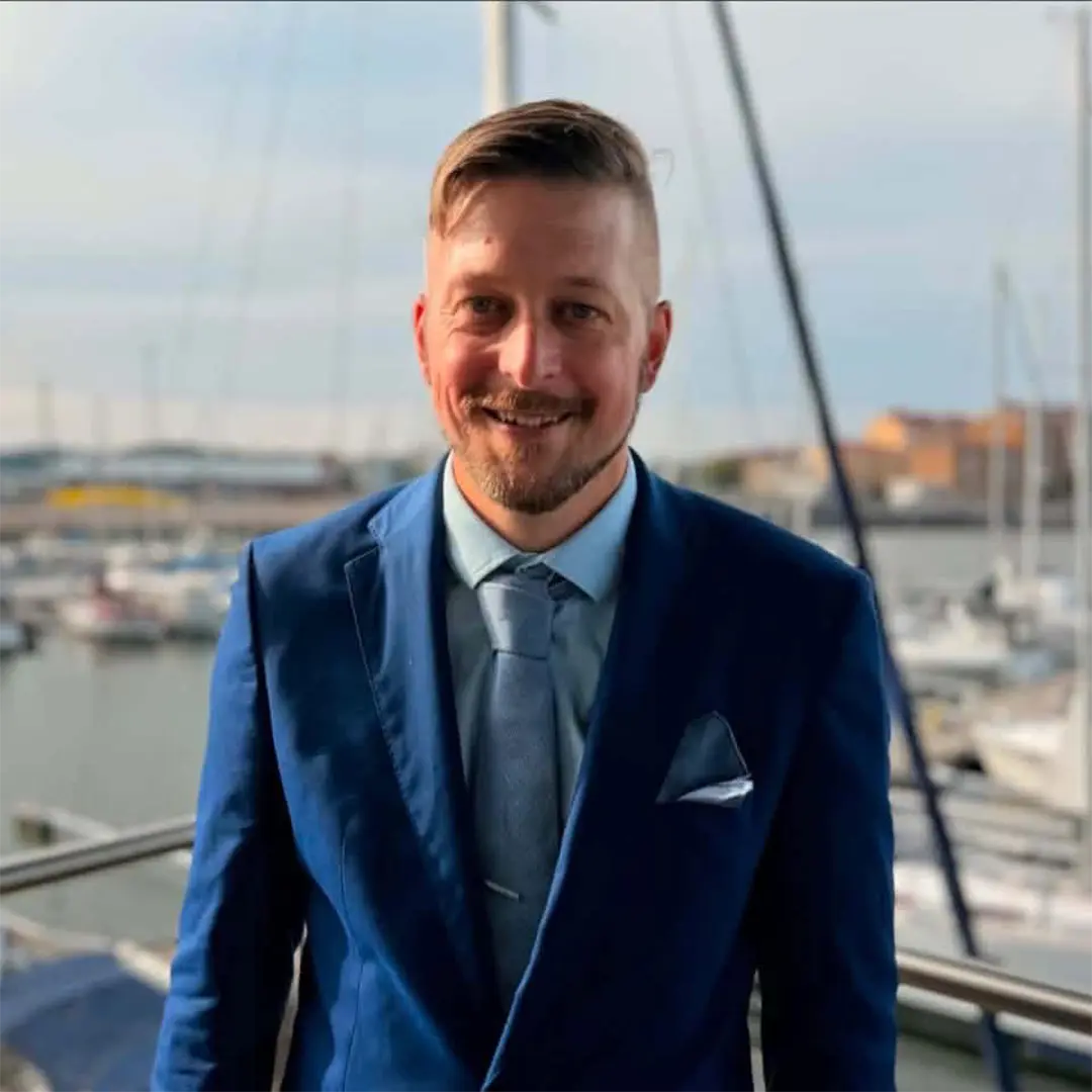 Porträttbild Johannes som studerar till platschef. Han är klädd i en kostym. Bakom honom finns en hamn med båtar.