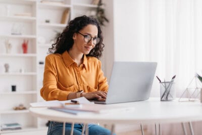 Kvinna med mörkt lockigt hår och glasögon, iklädd orange skjorta, sitter framför datorn vid ett skrivbord och jobbar digitalt.
